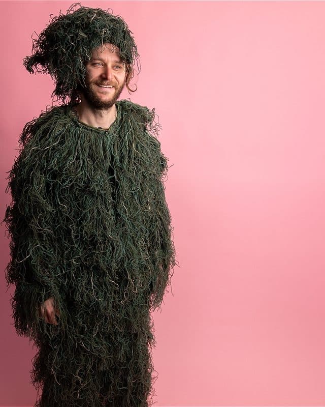 Mort is dressed in a ghillie suit with his face exposed, revealing a content smile.