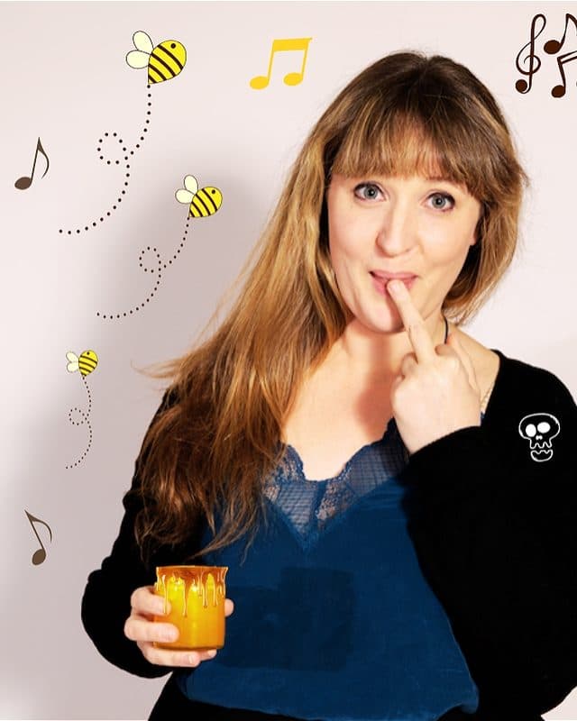 Anna PC dips her middle finger into a jar of honey, whilst smiling cheekily. In the background are music notes and bees.