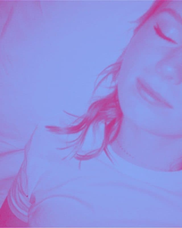 A close up of a young woman wearing a white top, with her eyes closed. She has light skin and red hair. Her face is half in the photo and half outside the frame, and there is a purple tint over the entire image, giving it a dreamy quality.