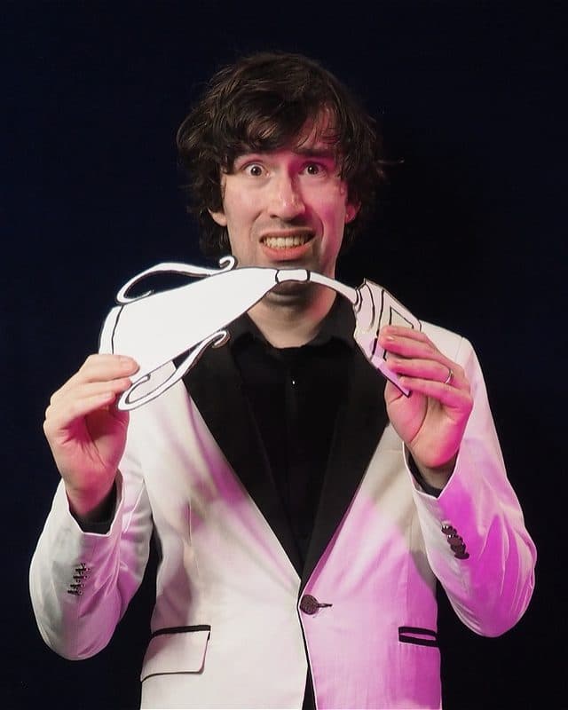 A Caucasian male in a white suit stands in front of a black backdrop. He is scared with wise brown eyes. He has messy dark brown hair. He holds a fake paper trophy he is bending.
