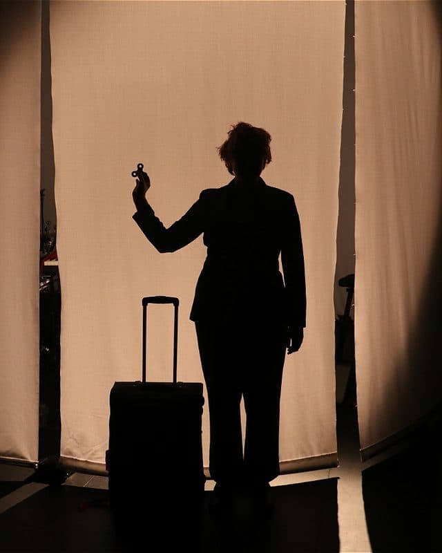 A silhouetted figure stands beside a carry-on suitcase, raising a fidget spinner against a glowing backlit curtain. The figure’s posture and formal attire evoke the feel of a cinematic travel campaign, while the warm lighting and deep shadows create a mysterious mood.