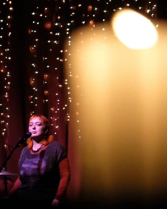 Madam Nightingale on stage about to sing into microphone in front of stage fairy lights with blurred image of candle in RHS foreground