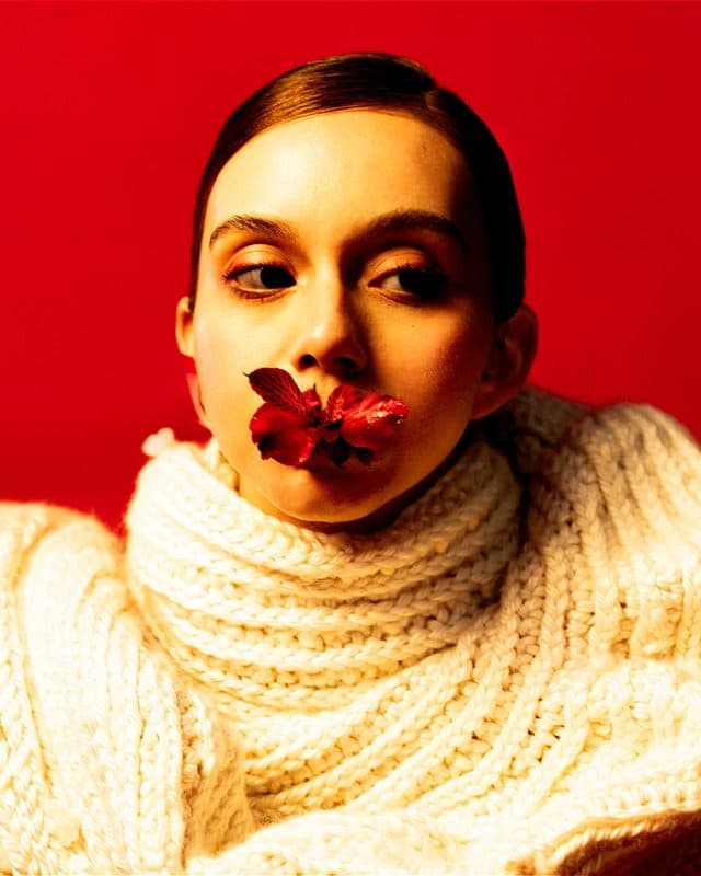 Woman in front of a red background wearing a white sweater. She has a red flower in her mouth and is looking to the left.