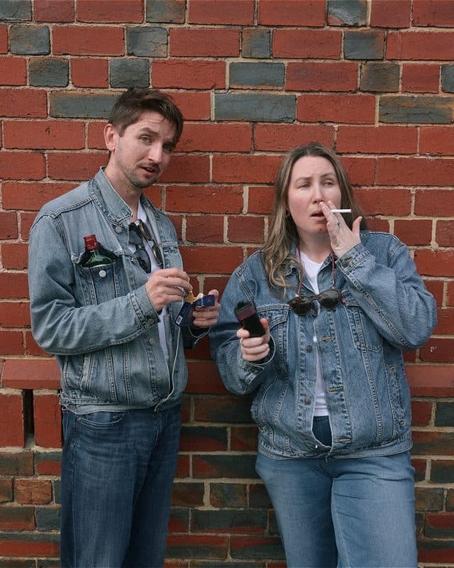 An image of Dan Hough and Tamara Absalom wearing denim jackets and jeans, ironically holding unlit cigarettes and holding bottles of alcohol in front of a brick wall.