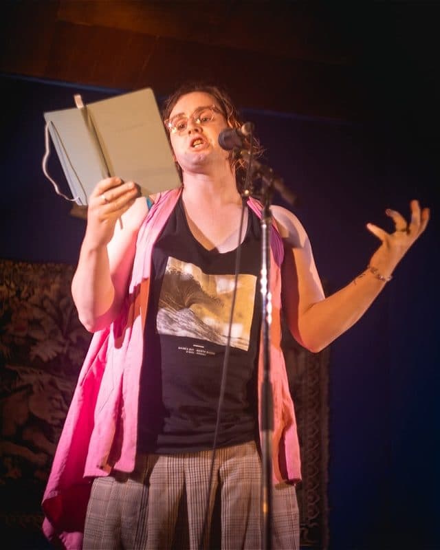 Alexi stands alone on a dimly lit stage performing at a floor microphone, holding an open notebook. She wears a black graphic singlet, grey pleated skirt, and a bright pink sleeveless vest draped over her shoulders. The background is a deep blue wall hung with framed paintings and a tapestry, with a warm floor lamp glowing to her left.