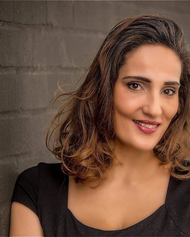 Portrait of Suha against a brick background, wearing a black top and smiling softly at the camera.