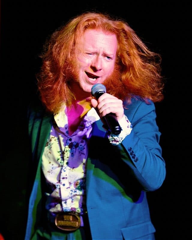 Host of The Doghouse LIVE, Corgi. A red haired male presenting individual in front of a black background with flowing red hair, wearing a bright blue suit, gripping a microphone with a passionate expression